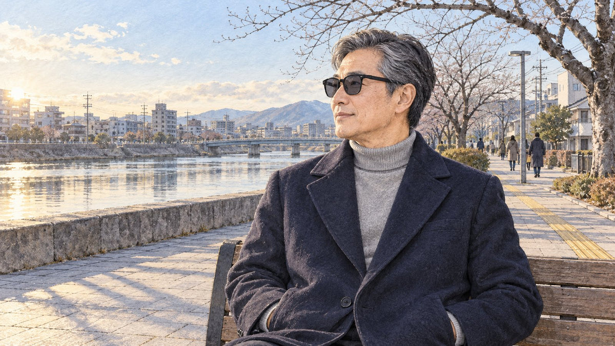 冬の川沿いのベンチに座り、コートを着てサングラスをかけた男性